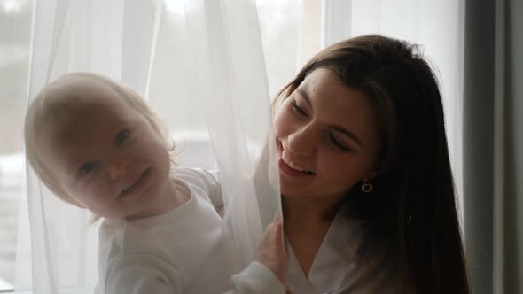 a Mother in a White Coat Sits with Her Daughter in a Bodysuit at a Large Window