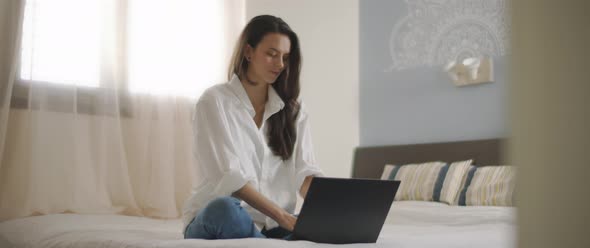 A woman in white shirt working on a computer, while sitting in bed