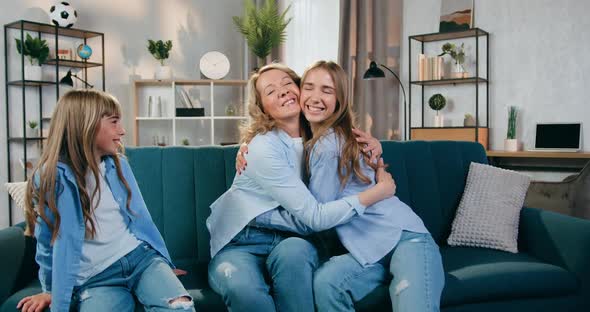 Positive Adult Mom Hugging Her Elderst and Younger Daughters Sitting with them on the Sofa at Home