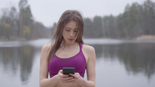 Portrait of Charming Young Caucasian Woman in Violet Sportswear Using Smartphone and Smiling