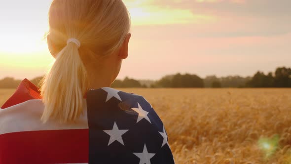 A Woman with a USA Flag on Her Shoulders Enjoys a Field of Wheat at Sunset