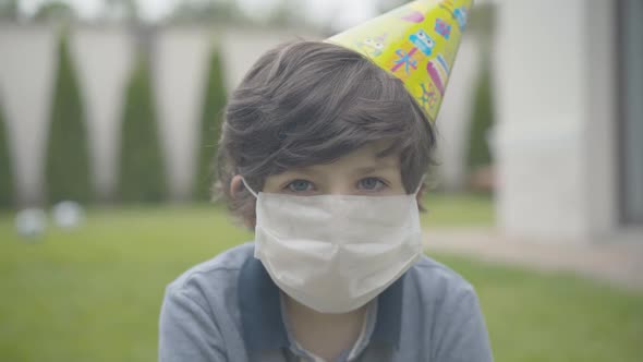 Close-up of Sad Caucasian Brunette Boy in Face Mask and Party Hat Looking at Camera. Portrait of