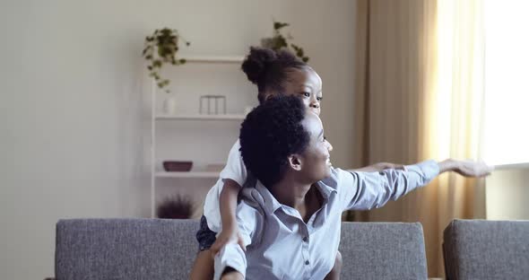 Afro American Black Woman Mom with Daughter Pretend to Be Airplane While Sitting on Sofa in Living