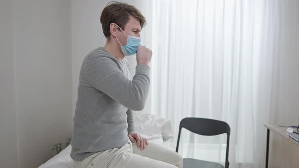 Side View Portrait of Sick Caucasian Man in Covid19 Face Mask Sitting on Medical Couch Coughing