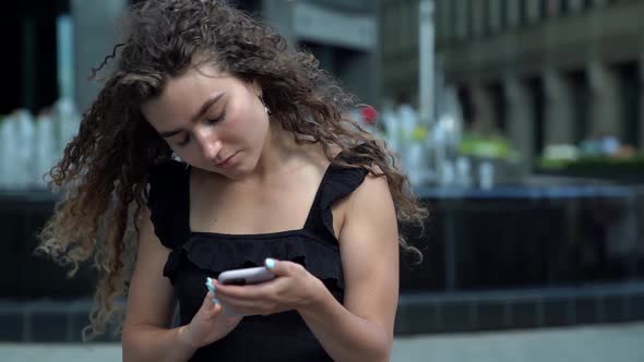 Portrait of a Young Woman with Curly Hair and a Phone in Her Hands on the Background of a Fountain