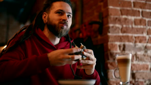 Portrait of Trendy Guy with Dreadlocks Eating Burger in Fast Food Restaurant Medium Shot