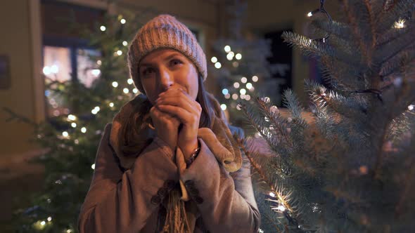 Portrait of a Happy Woman Against the Backdrop of Christmas Decorations. She Rubs Her Hands, Keeping