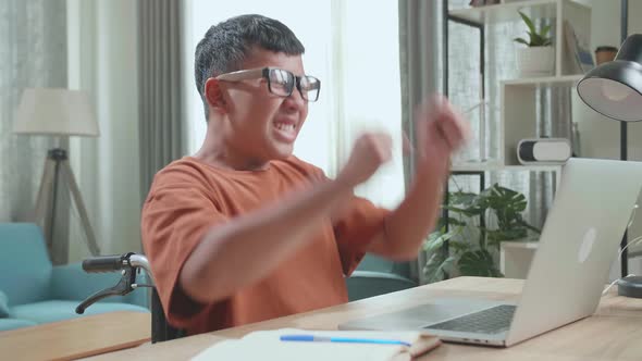 Young Asian Boy Sitting In A Wheelchair While Using Laptop Computer And Celebrating At Home