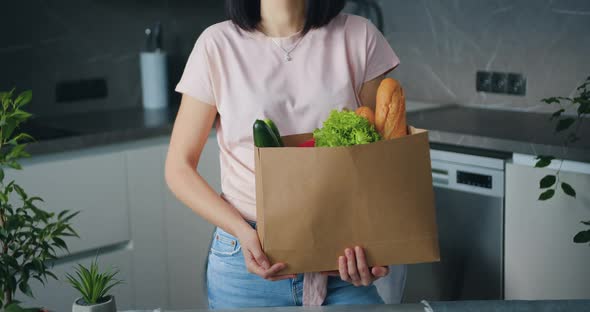 Woman with Dark Hair Looking at Camera in the Kitchen Holding Paper Package with Food 