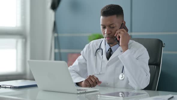 African Doctor Talking on Smartphone while using Laptop in Office