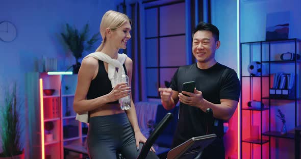 Asian Guy Talking with His Pretty Fit Athletic Blond Girlfriend which Exercising on Trainer Bicycle