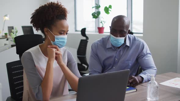 Diverse businessman and businesswoman in face masks discussing, using laptop in office