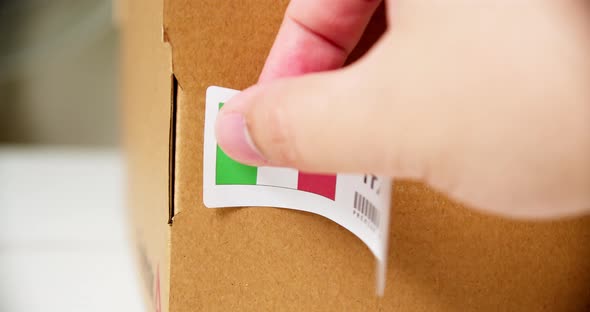 Hands applying MADE IN ITALY flag label on a shipping cardboard box with products. Close up shot wit