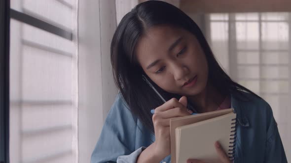 Young Asian businesswoman standing beside window talking on the phone and taking notes.