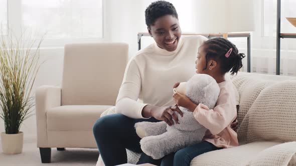 Happy African Mother with Afro Little Daughter Child Schoolgirl Sitting on Sofa at Home Talking