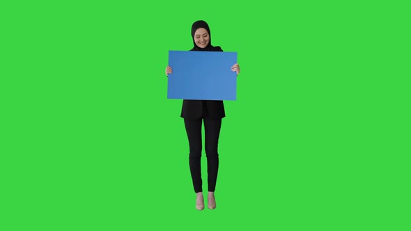 Smiling Arab Woman in Hijab Holding Blank Blue Poster and Looking at It on a Green Screen, Chroma