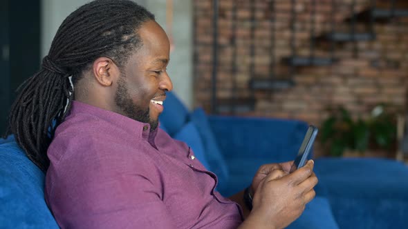 Smiling AfricanAmerican Man Using Smartphone Sitting on the Sofa at Home