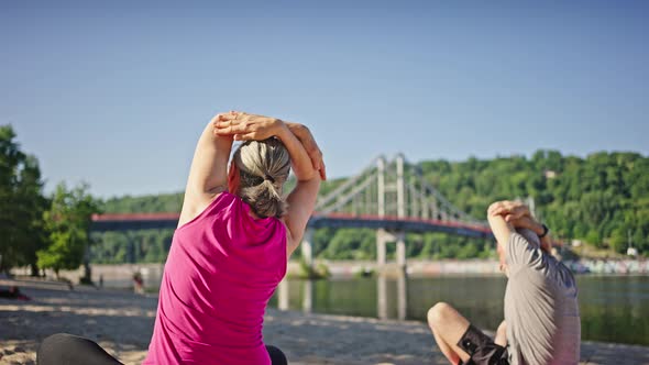 Greyhaired Pensioners Stretch on Sand Against Bridge