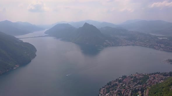 Aerial drone shot over the forest and a large misty blue lake next to the town buildings in Lugano,