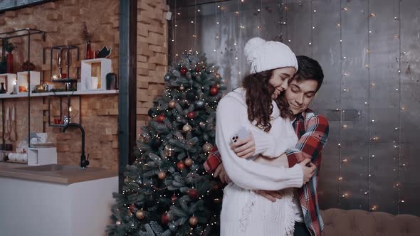 Young Couple Embracing at Home Near the Christmas Tree, Using Their Mobile Phone To Track the New