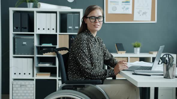 Portrait of Cheerful Young Lady in Wheelchair Smiling Looking at Camera Indoors in Office