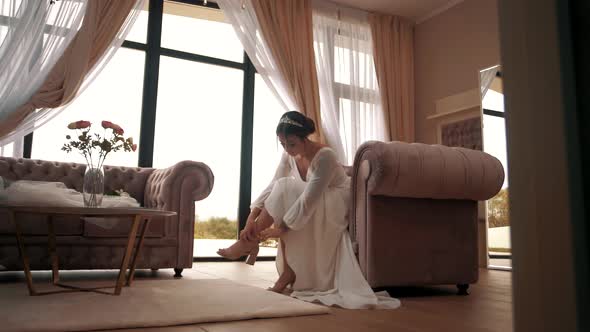 Young bride sits in an armchair and elegantly puts on her wedding shoes. General wide shot