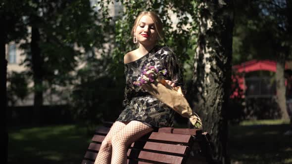 Young Slim Annoyed Drunk Woman Smelling Flowers Dropping Bouquet Sitting on Bench in City Park in