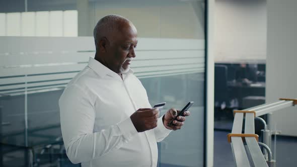 Mature Black Businessman Using Credit Card And Smartphone At Workplace In Office