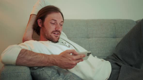 Concentrated long-haired man watching video on phone while lying on the sofa