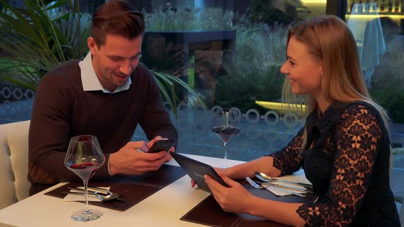 A Man and a Woman Sit at a Table in a Restaurant and Work on Smartphone and Tablet and Talk