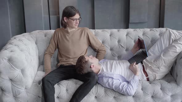 Young Positive Man Lying on Couch with Laptop and Talking with Male Partner. Caucasian Gay Couple