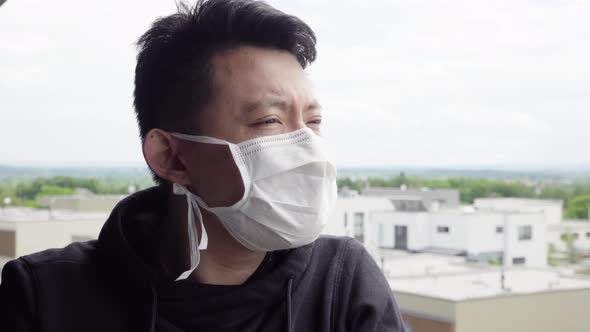 A Young Asian Man in a Face Mask Stands on a Balcony and Looks at a Cityscape Below