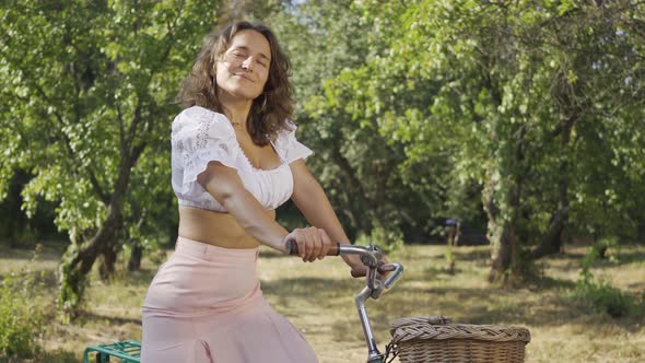 Cute Young Woman with Curly Hair Standing in the Garden or Park with Her Bicycle