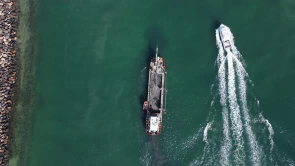 A luxury boat passing a large industrial sand dredging ship next to a coastal seaway rock wall. High