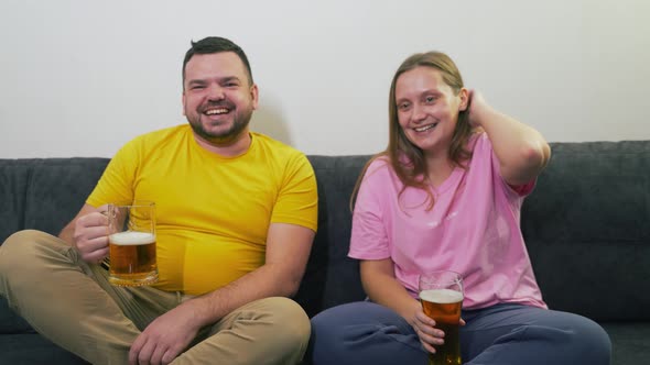 Young Couple are Sitting on Gray Sofa Watching TV and Drinking Beer From Transparent Glass Glasses