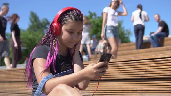 Girl Listening to Music on Headphones Using Smartphone Sitting on Bench in Summer Park