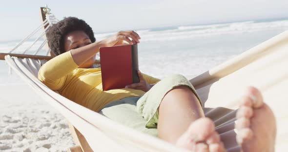 Happy african american woman reading and lying in hammock on sunny beach