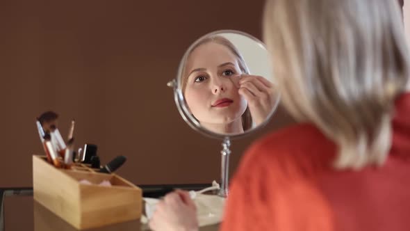 Beautiful blond hair woman in red shirt applying makeup in opposite of mirror