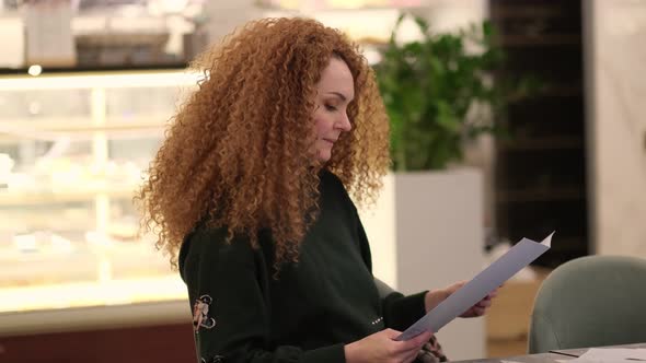 Bright Redhead Woman Sits at a Table in a Cafe and Chooses a Dish From the Menu