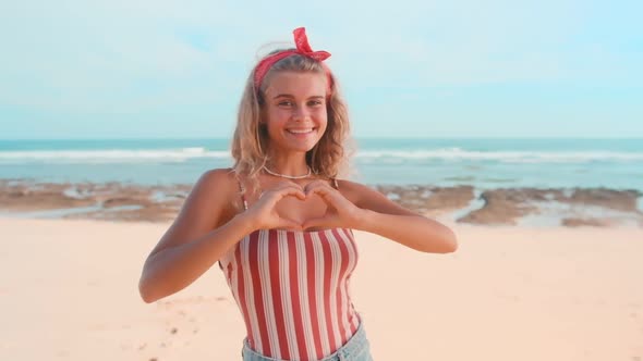 Young Caucasian Woman Zoomer with Heart Made of Fingers Stands on Sunny Beach