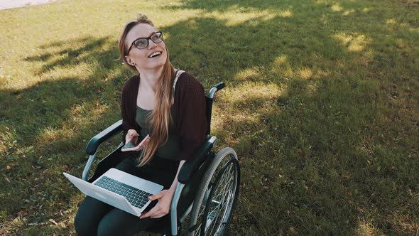 Happy Young Woman in Wheelchair with Laptop and Smartphone Looking Up in the Sky.