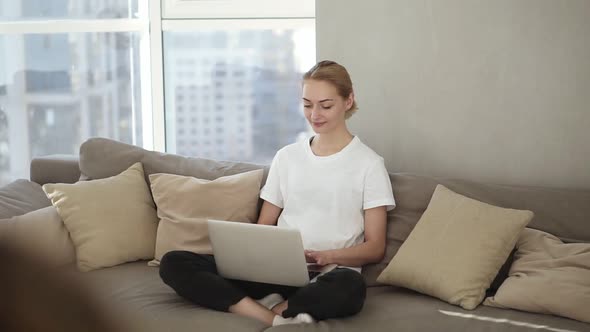 Smiling Woman Freelancer Sitting on Couch in Domestic Clothes Freelance From Home Typing Email on