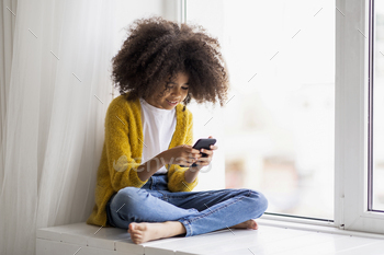 g on windowsill, using cell phone at home, happy african american kid chatting with her friends or using newest mobile app, copy space