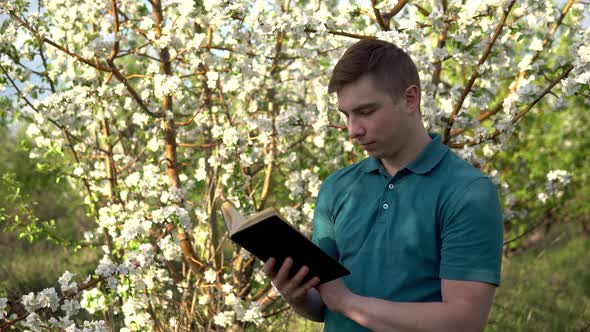 Young Man on Nature with a Book in His Hands. A Man Reads a Book While Standing Against a Flowering