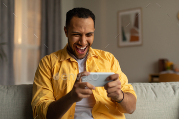 excited over winning lottery or casino bet online, sitting on couch at home. Emotional African American guy playing video game, gambling indoors