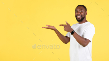 Hand Holding Invisible Object Smiling To Camera Standing Over Yellow Background. Man Advertising Product In Studio. Panorama