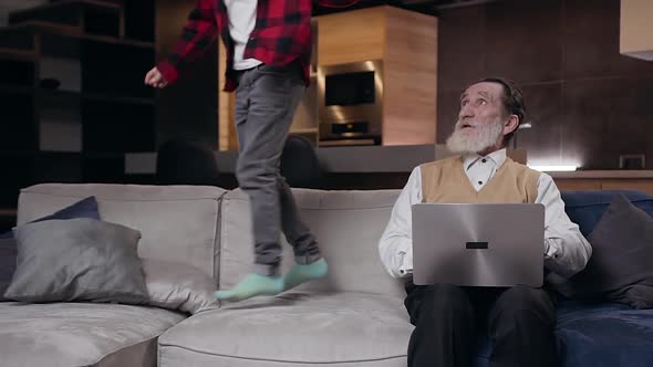 Boy which Jumping Near His Serious Mature Bearded Grandfather and Interfering Him while He Working