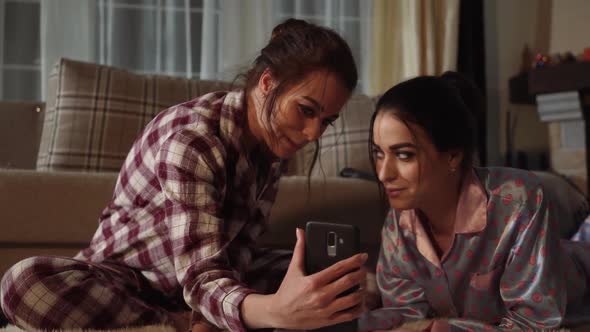 Two Sisters Twins Watching Something Funny on Smartphone and Laughing on the Background of Cozy