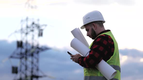 Worker Male Engineer Using Mobile Phone for Checking Data While Standing Against High Voltage Power