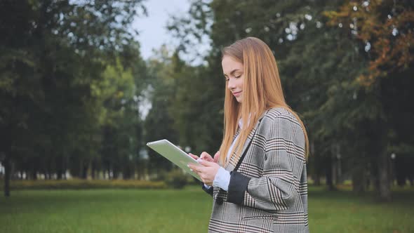 A Young Girl Walks with a Tablet in the Park